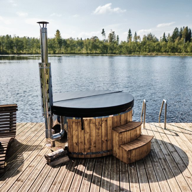 Holzbeheizter Outdoor-Hot-Tub mit schwarzer Abdeckung auf einem Steg an einem See, umgeben von Wald und klarem Himmel.