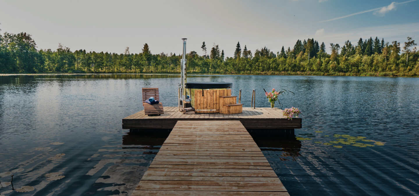 Holzbadetonne auf einem Steg mitten auf einem ruhigen See, umgeben von Bäumen und Natur.
