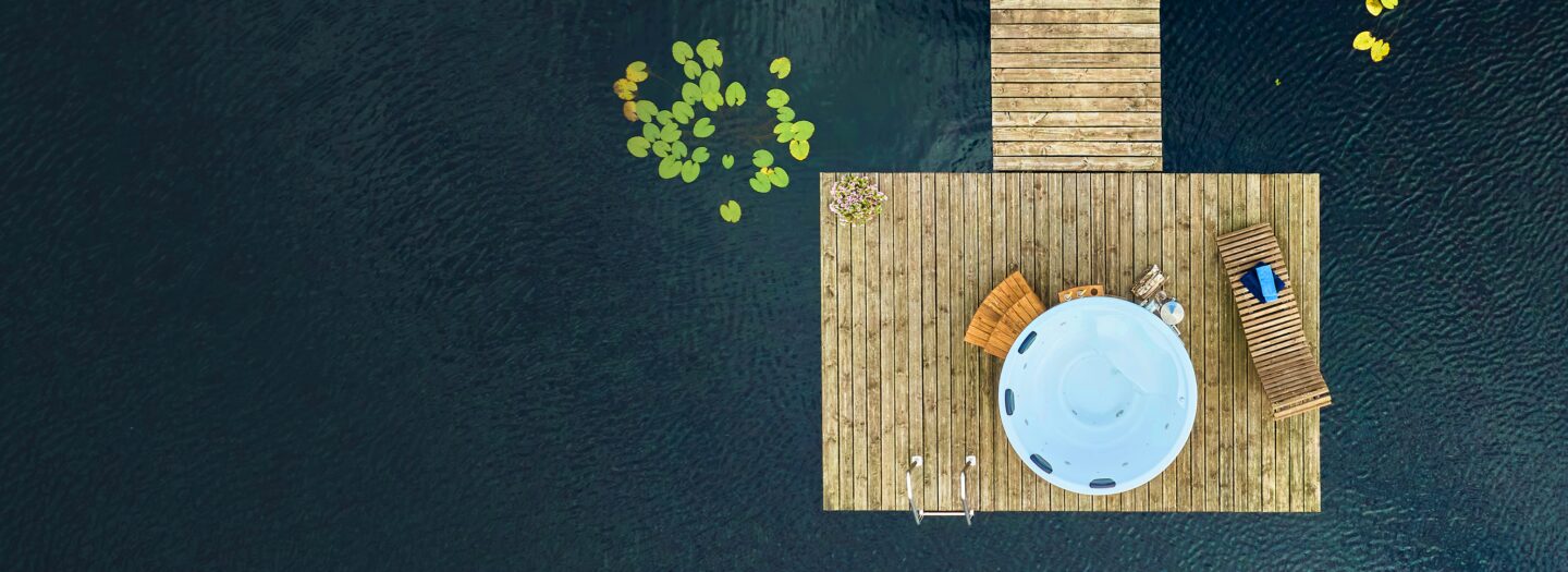 Holzbefeuerter Hot Tub von Feuerbad mit Holztreppe auf einem Steg über einem See aus der Vogelperspektive. Neben dem Hot Tub steht eine Liege mit zusammengerollten Handtüchern