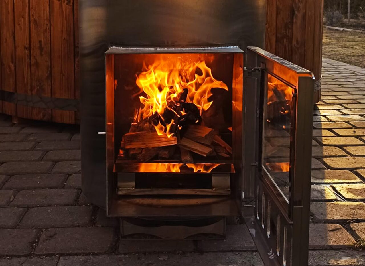 Dunkler Metall-Ofen an einem Feuerbad Hot Tub mit geöffneter Tür. Brennendes Holz ist zu sehen.