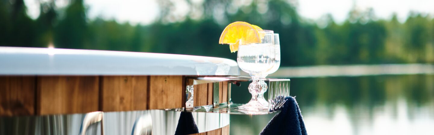 Zu sehen ist ein Hot Tub von Feuerbad. Auf einer Ablage stehen 2 Gläser und es hängt ein dunkles Handtuch daran. Im Hintergrund sieht man einen von Bäumen umgebenen See