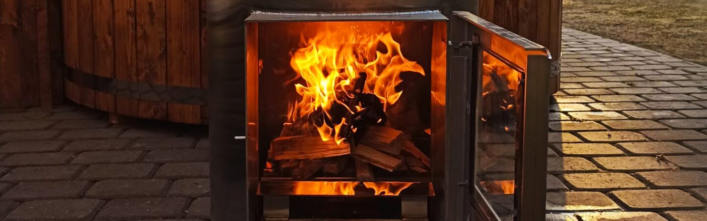 Zu sehen ist, ein Metallofen mit geöffneter Tür, in dem ein Feuer über aufgeschichteten Holzscheiten brennt. Der Ofen steht auf einem gepflasterten Außenbereich vor einem Hot Tub.