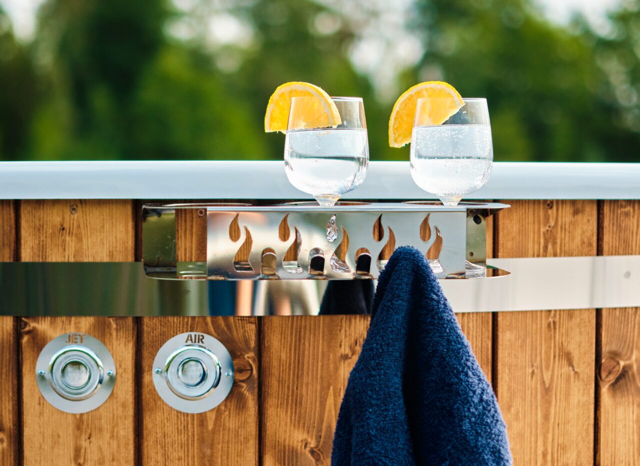 Zu sehen ist ein Hot Tub von Feuerbad. Auf einer Ablage stehen 2 Gläser und es hängt ein dunkles Handtuch daran. Im Hintergrund sieht man Bäume